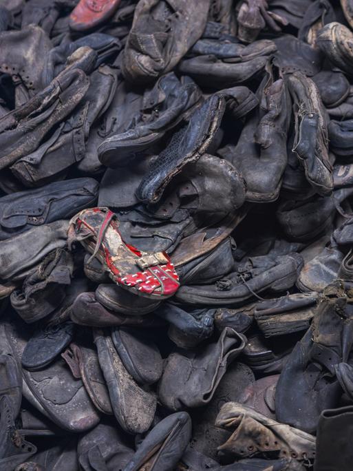 Unzählige dunkle Schuhe auf einem Haufen, ein roter Schuh liegt oben drauf. Es sind Schuhe in der Gedenkstätte Auschwitz-Birkenau.