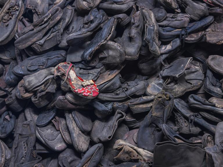 Unzählige dunkle Schuhe auf einem Haufen, ein roter Schuh liegt oben drauf. Es sind Schuhe in der Gedenkstätte Auschwitz-Birkenau.