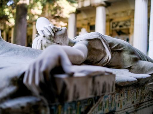 Statue eines liegenden Engels, der sich eine Hand vors Gesicht hält, auf einem Grab