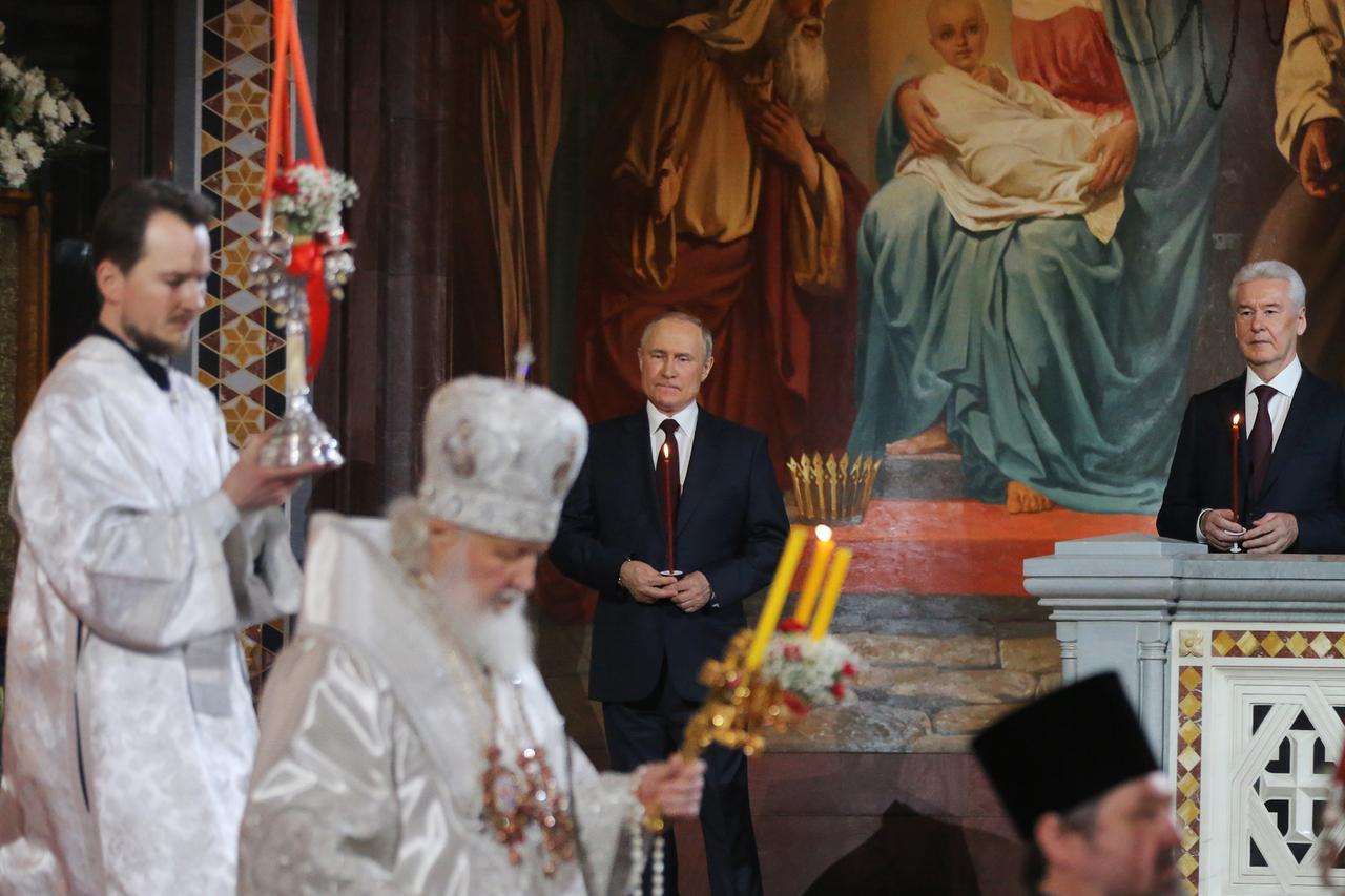 Wladimir Putin steht im Bildhintergrund und hält eine Kerze in den Händen. Im Bildvordergrund ist das Oberhaupt der russisch-orthodoxen Kirche, Kyrill,  ebenfalls mit Kerzen, zu sehen.