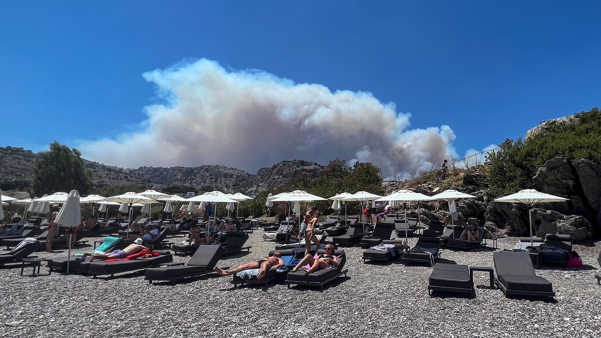 Der Rauch der Waldbrände erhebt sich der Urlaubsinsel Rhodos auf, während im Vordergrund Urlauber auf ihren Sonnenliegen entspannen.