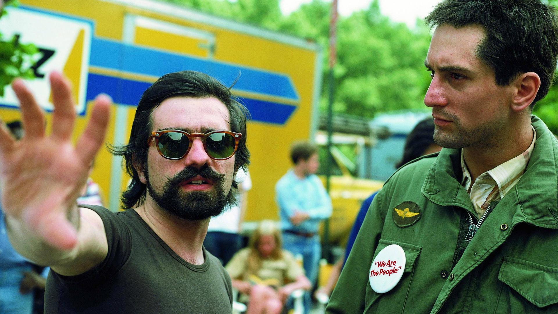 Martin Scorsese trägt eine Sonnenbrile und gibt Robert De Niro gestikulierend Regieanweisungen. Beide stehen im Freien.