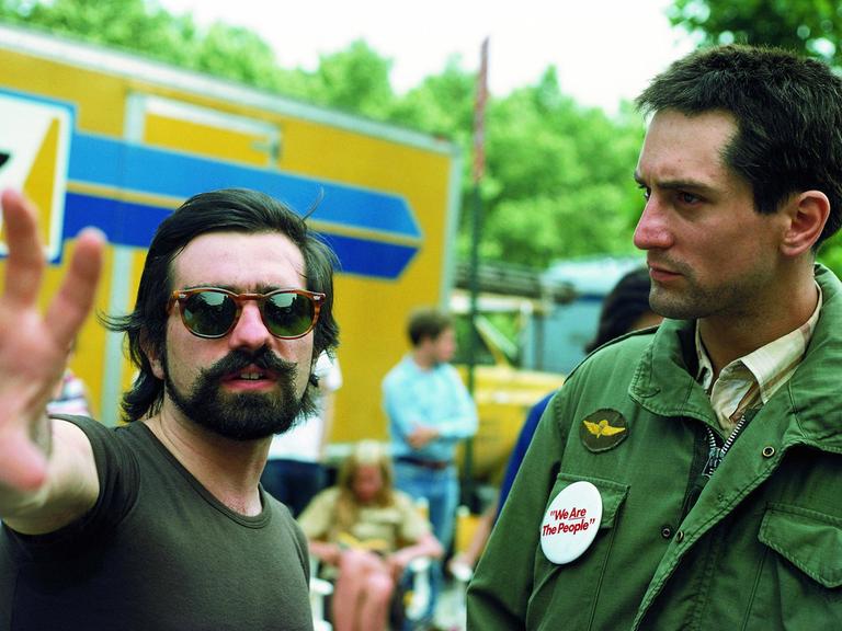 Martin Scorsese trägt eine Sonnenbrile und gibt Robert De Niro gestikulierend Regieanweisungen. Beide stehen im Freien.