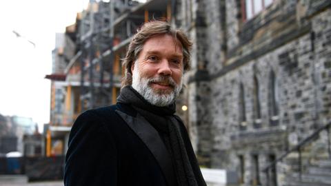Ein Mann mit braunen Haaren und grauem Vollbart blickt freundlich in die Kamera. Er steht vor einem großen Gebäude, das nur unscharf zu erkennen ist. Er trägt eine schwarze Anzugjacke und einen schwarzen Schal.