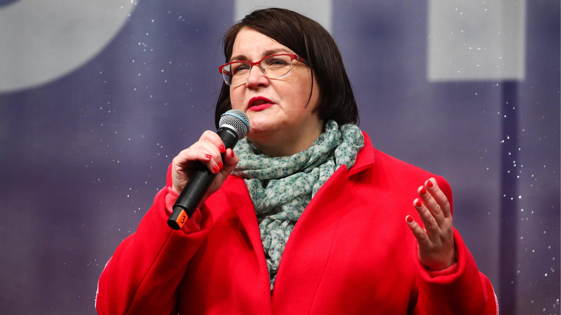 Das Bild zeigt Julia Galjamina auf einer Bühne mit einem Mikrofon in der rechten Hand. Sie trägt eine Brille und eine rote Jacke.