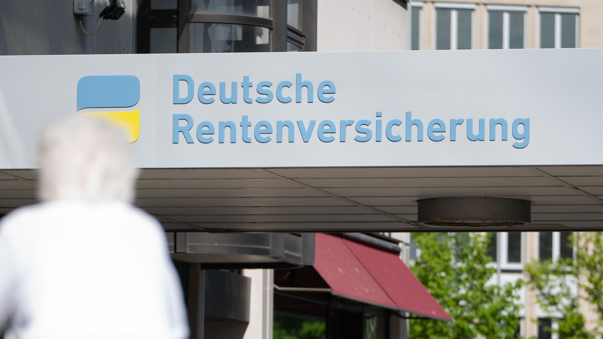 Eine Frau mit weißen Haaren läuft vor einem Haus mit dem Logo der Deutsche Rentenversicherung entlang. Eine Frau mit weißen Haaren läuft vor einem Haus mit dem Logo der Deutsche Rentenversicherung entlang.