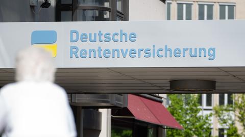 Eine Frau mit weißen Haaren läuft vor einem Haus mit dem Logo der Deutsche Rentenversicherung entlang. 