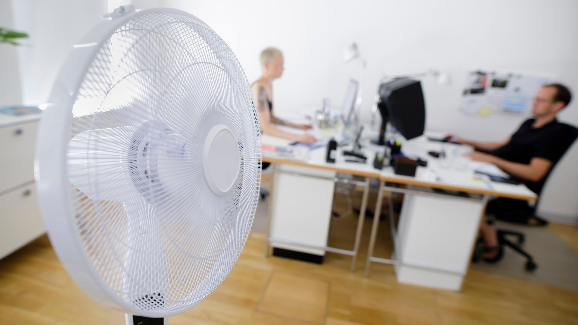 Ein Ventilator dreht steht in einem Büro. Im Hintergrund sitzen zwei Mitarbeitende sich an ihren Schreibtischen gegenüber.