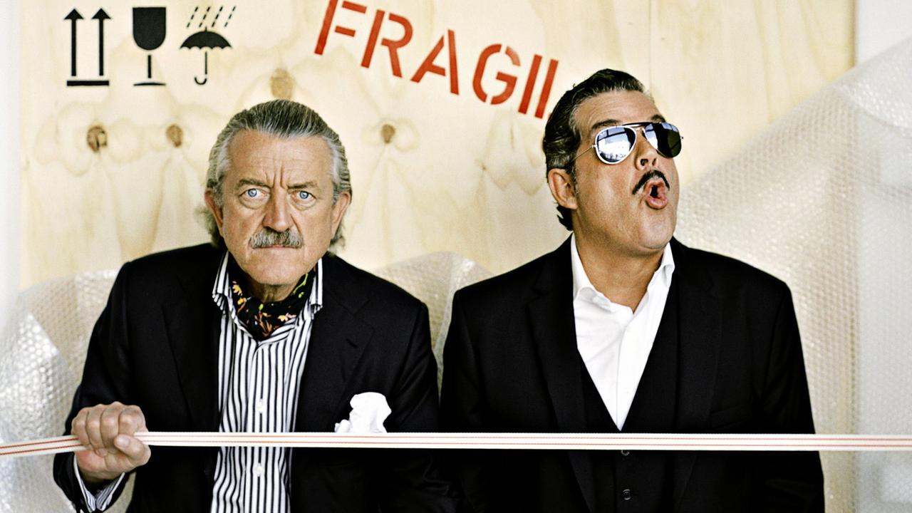 Dieter Meier und Boris Blank von der Band Yello posieren in schwarzen Anzügen vor einer Holzwand mit der Aufschrift "Fragil".