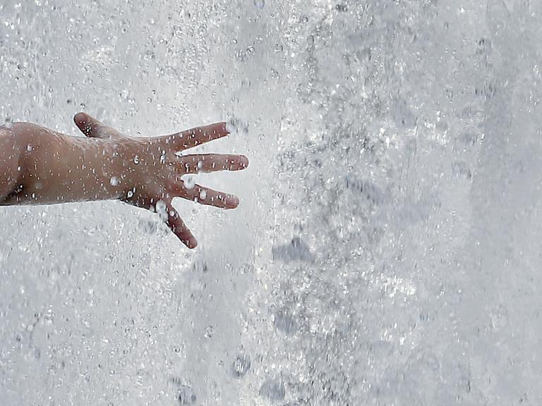 Das Bild zeigt eine Kinderhand, die bei bei Temperaturen um 31 Grad Celsius in eine Springbrunnenanlage mit sprudelndesm Wasser greift.