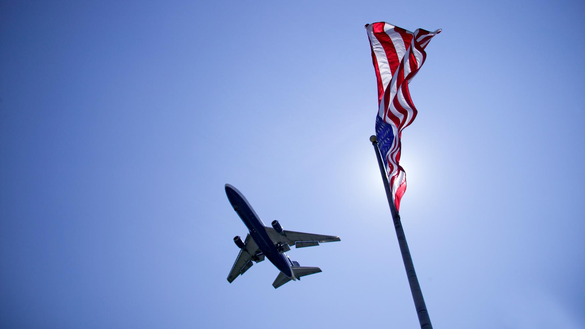 Untersicht auf ein Flugzeug am Himmel. Rechts im Bild die amerikanische Flagge.