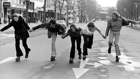 Fünf Menschen halten sich an den Händen und fahren Rollschuh auf einer leeren Straße.