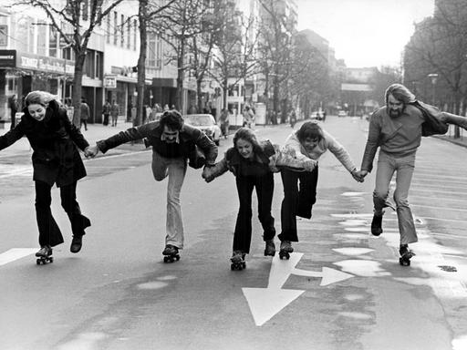 Fünf Menschen halten sich an den Händen und fahren Rollschuh auf einer leeren Straße.