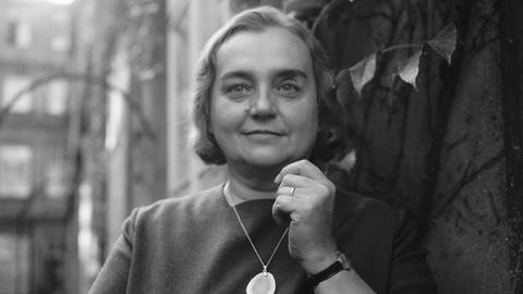 Historisches Schwarzweißporträt von Christine Busta, die vor einer Hauswand steht und in die Kamera lächelt. Ihr kurzes Haar liegt in Wellen und um den Hals trägt sie eine Kette mit einem großen Halbedelstein.