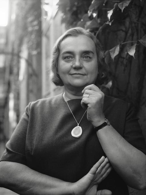 Historisches Schwarzweißporträt von Christine Busta, die vor einer Hauswand steht und in die Kamera lächelt. Ihr kurzes Haar liegt in Wellen und um den Hals trägt sie eine Kette mit einem großen Halbedelstein. Historisches Schwarzweißporträt von Christine Busta, die vor einer Hauswand steht und in die Kamera lächelt. Ihr kurzes Haar liegt in Wellen und um den Hals trägt sie eine Kette mit einem großen Halbedelstein.