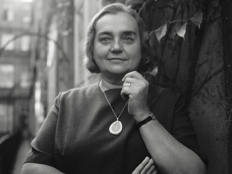 Historisches Schwarzweißporträt von Christine Busta, die vor einer Hauswand steht und in die Kamera lächelt. Ihr kurzes Haar liegt in Wellen und um den Hals trägt sie eine Kette mit einem großen Halbedelstein. Historisches Schwarzweißporträt von Christine Busta, die vor einer Hauswand steht und in die Kamera lächelt. Ihr kurzes Haar liegt in Wellen und um den Hals trägt sie eine Kette mit einem großen Halbedelstein.