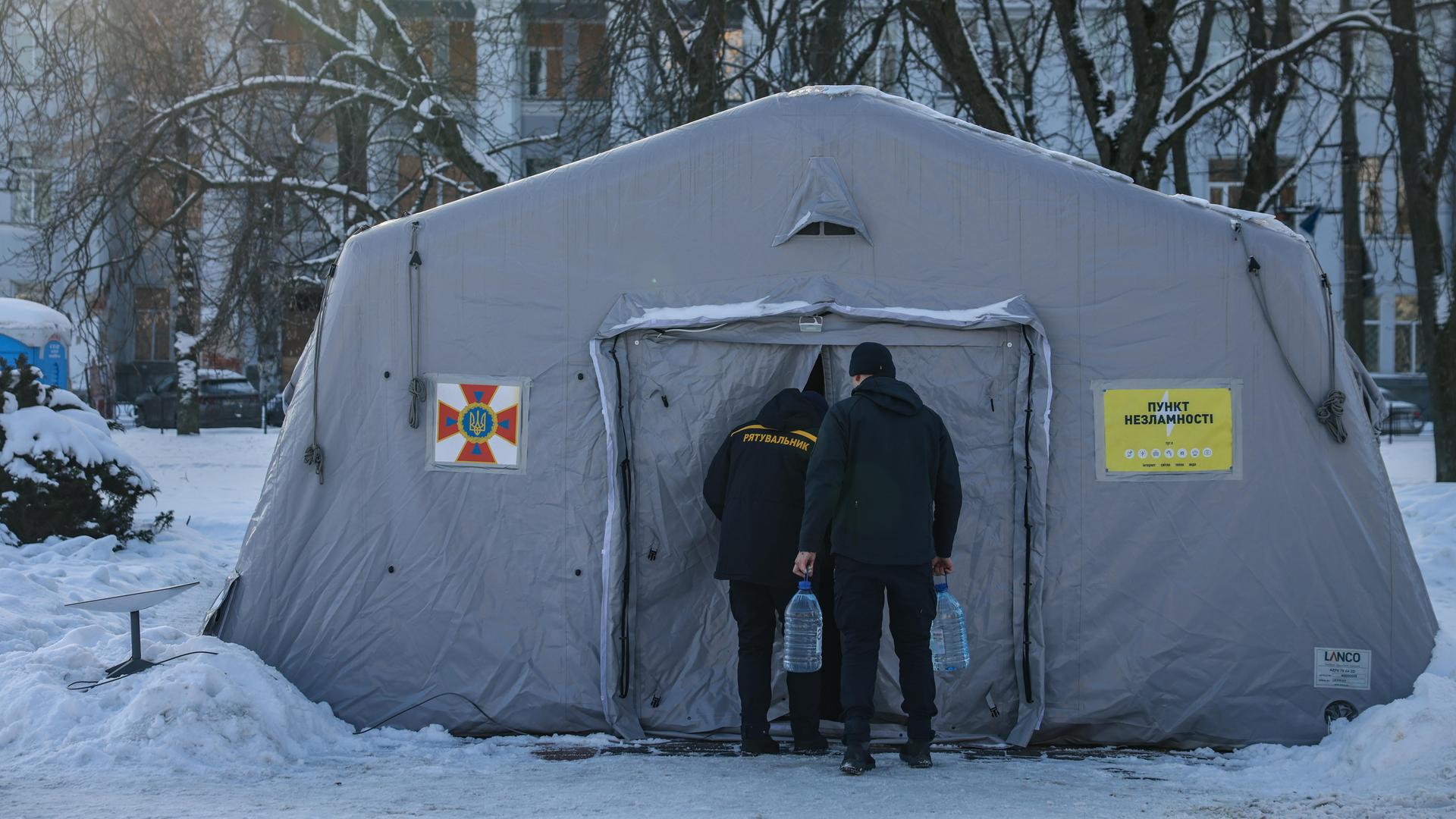 Zwei Männer gehen in ein großes, weißes Heizzelt, das von Schnee und Wohnhäusern umgeben ist.