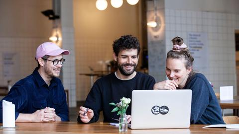 An einem Tisch in einem großen halb-gefliesten Raum sitzen drei Menschen und schauen auf einen Laptop.