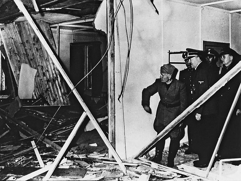 Benito Mussolini und Adolf Hitler, begleitet von weiteren Männern in Uniform, schauen in einen von Trümmern übersäten Raum.
