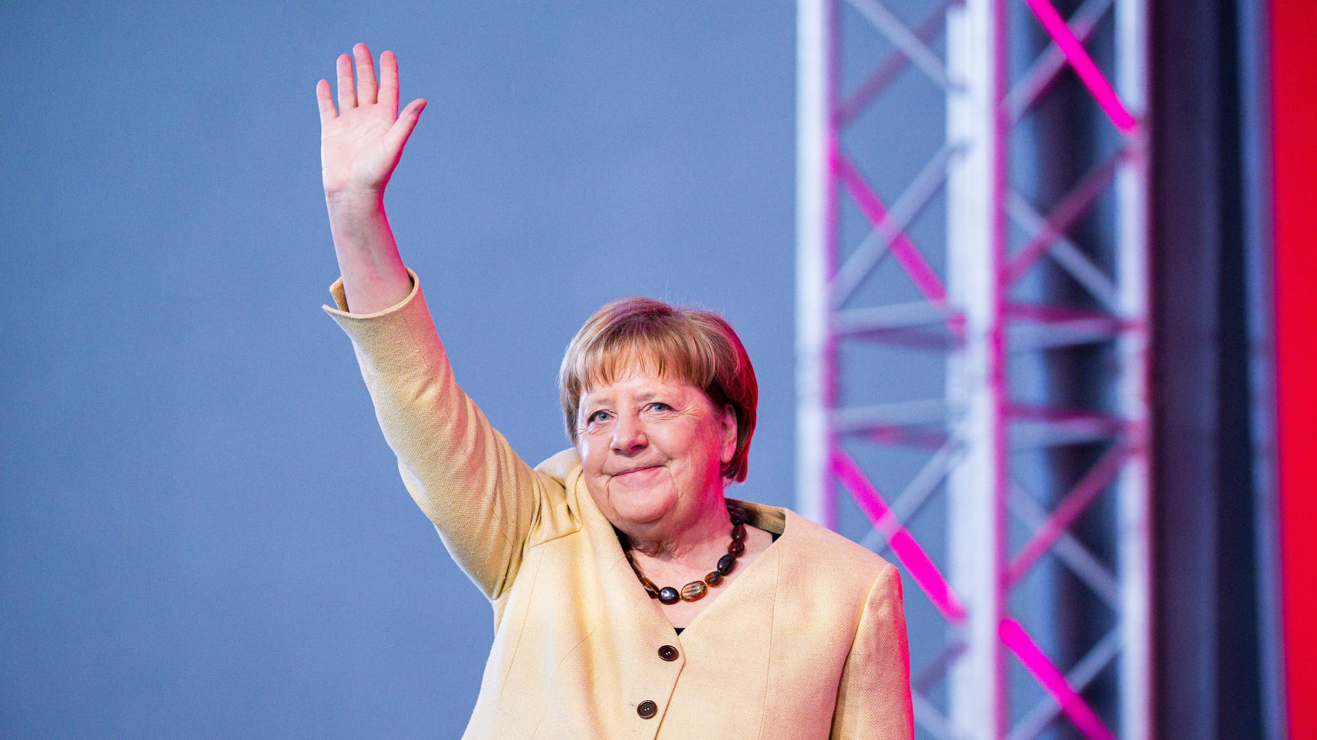 Das Bild zeigt die ehemalige deutsche Kanzlerin Angela Merkel winkend auf einer Bühne beim Deutschen Evangelischen Kirchentag am 1. Mai 2025. Sie lächelt ins Publikum und trägt eine hellbeige Anzugjacke sowie eine bernsteinfarbene Kette. Das Bild zeigt die ehemalige deutsche Kanzlerin Angela Merkel winkend auf einer Bühne beim Deutschen Evangelischen Kirchentag am 1. Mai 2025. Sie lächelt ins Publikum und trägt eine hellbeige Anzugjacke sowie eine bernsteinfarbene Kette.