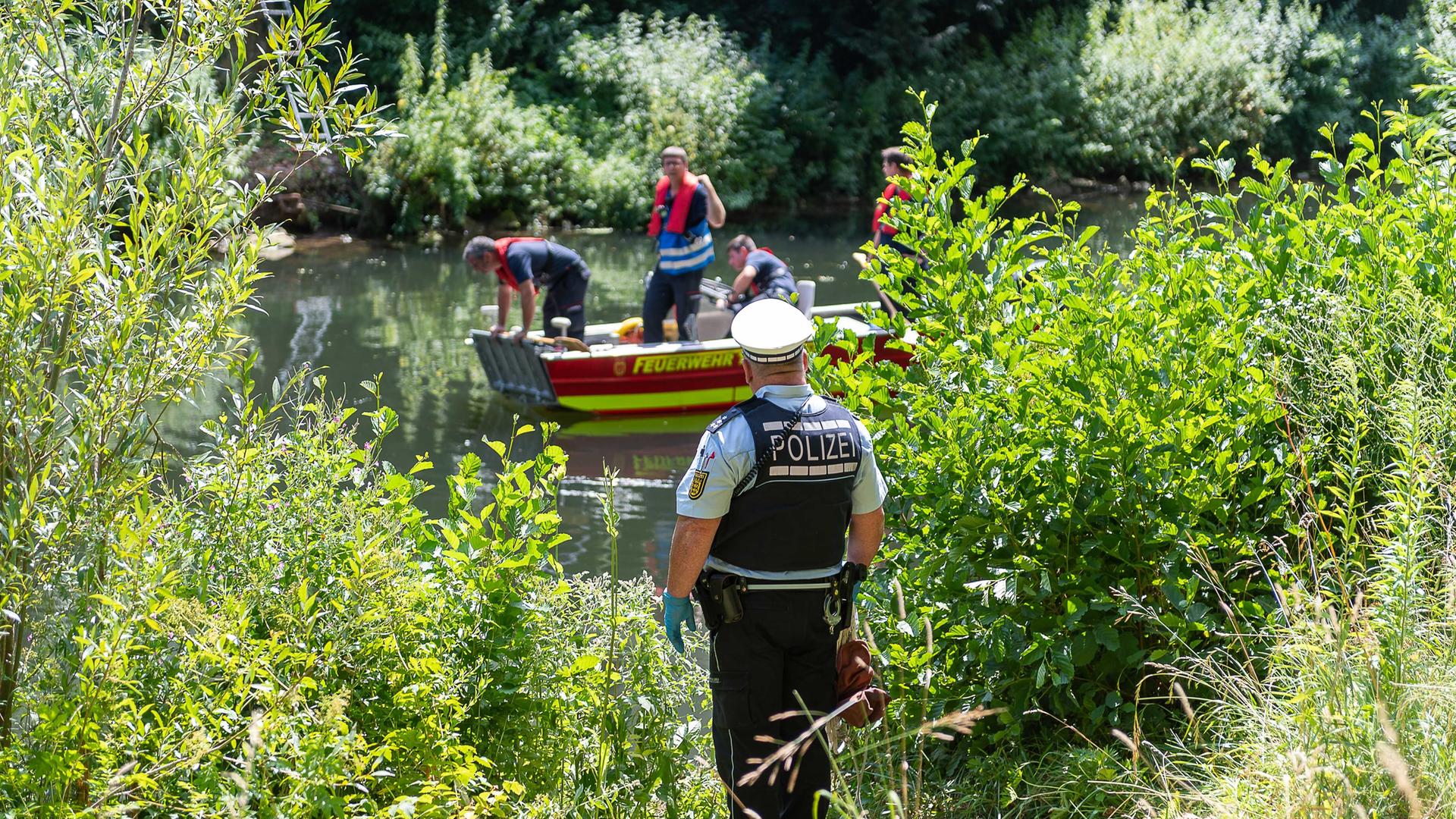 Feuerwehrleute und ein Polizist und suchen am Ufer eines Flusses und im Wasser nach einer Leiche.