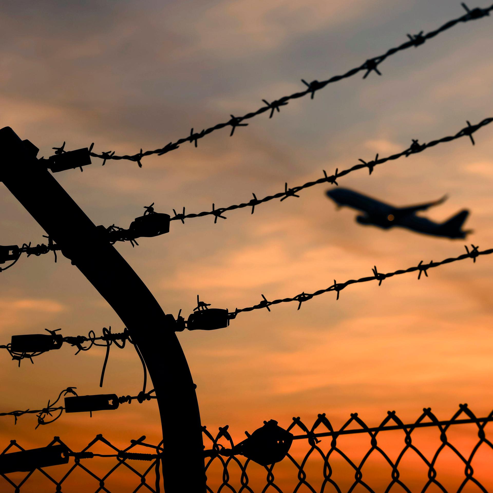 Ein Flugzeug startet am Flughafen Köln/Bonn, gesehen durch Stacheldraht außerhalb des Rollfelds. Es ist Sonnenuntergang.