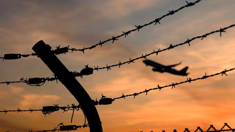Ein Flugzeug startet an einem Flughafen, gesehen durch Stacheldraht außerhalb des Rollfelds. Es ist Sonnenuntergang.