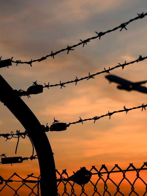 Ein Flugzeug startet am Flughafen Köln/Bonn, gesehen durch Stacheldraht außerhalb des Rollfelds. Es ist Sonnenuntergang.
