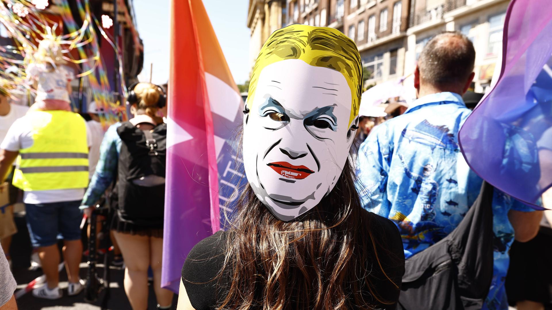 Eine Person auf einer Demonstration trägt eine weiße Pappmaske mit dem stilisierten Gesicht von Viktor Orbán. Die Maske zeigt ein streng wirkendes Gesicht mit zusammengezogenen Augenbrauen und rotem Mund. 