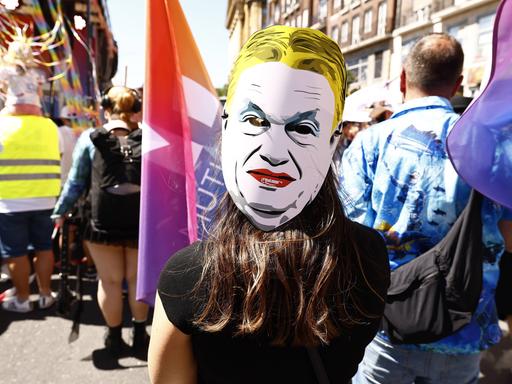Eine Person auf einer Demonstration trägt eine weiße Pappmaske mit dem stilisierten Gesicht von Viktor Orbán. Die Maske zeigt ein streng wirkendes Gesicht mit zusammengezogenen Augenbrauen und rotem Mund. 