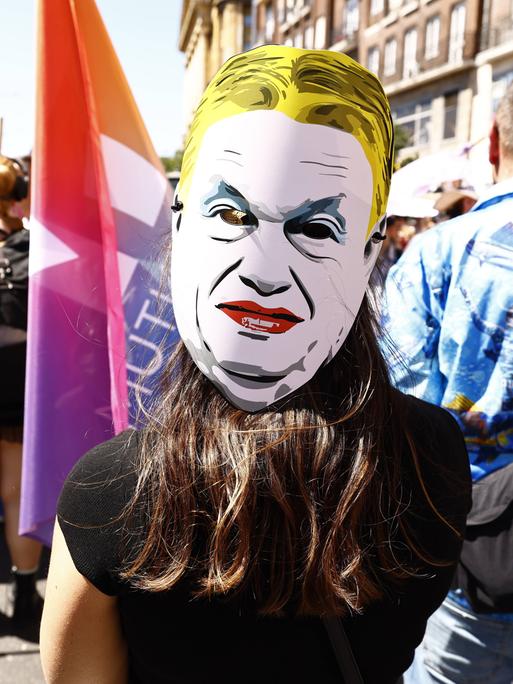 Eine Person auf einer Demonstration trägt eine weiße Pappmaske mit dem stilisierten Gesicht von Viktor Orbán. Die Maske zeigt ein streng wirkendes Gesicht mit zusammengezogenen Augenbrauen und rotem Mund. 