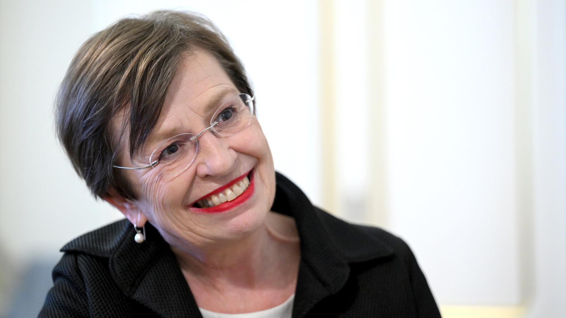 Doris Schmidauer in hellem Oberteil und schwarzem Kostüm mit Brille vor weißem Hintergrund.