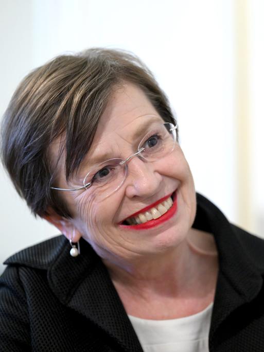 Doris Schmidauer in hellem Oberteil und schwarzem Kostüm mit Brille vor weißem Hintergrund.