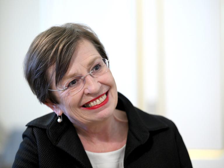 Doris Schmidauer in hellem Oberteil und schwarzem Kostüm mit Brille vor weißem Hintergrund.