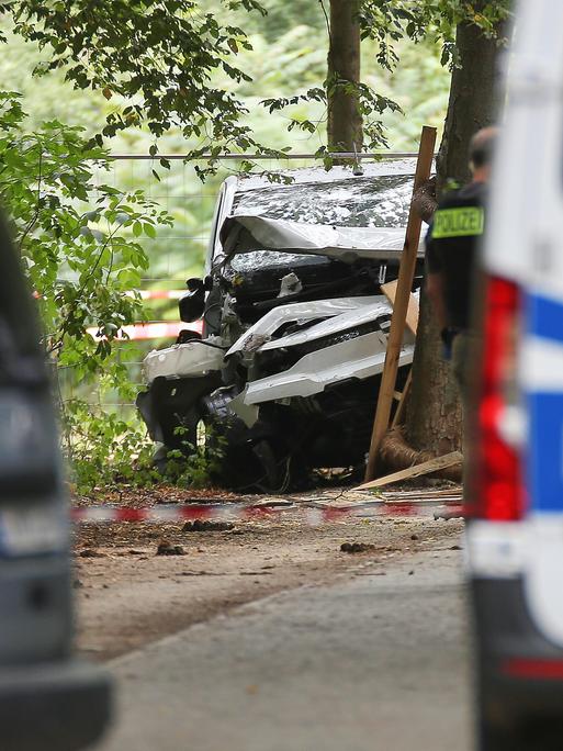 Zwischen zwei unscharf abgebildeten Polizeifahrzeugen ist in einem bewaldeten Bereich das stark beschädigte dunkle Tatfahrzeug zu sehen. Teile des Wagens sind eingedrückt, daneben steht eine Person in Polizeiweste. Zwischen zwei unscharf abgebildeten Polizeifahrzeugen ist in einem bewaldeten Bereich das stark beschädigte dunkle Tatfahrzeug zu sehen. Teile des Wagens sind eingedrückt, daneben steht eine Person in Polizeiweste.