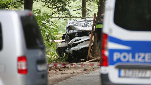 Zwischen zwei unscharf abgebildeten Polizeifahrzeugen ist in einem bewaldeten Bereich das stark beschädigte dunkle Tatfahrzeug zu sehen. Teile des Wagens sind eingedrückt, daneben steht eine Person in Polizeiweste.