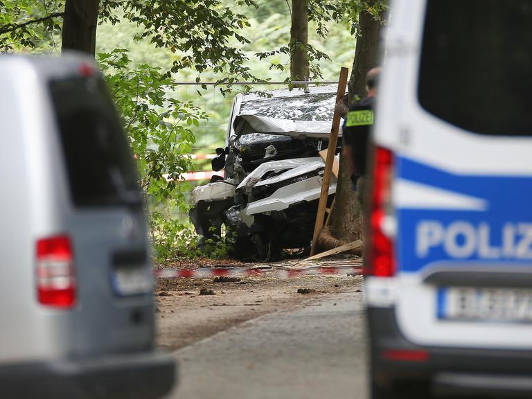 Zwischen zwei unscharf abgebildeten Polizeifahrzeugen ist in einem bewaldeten Bereich das stark beschädigte dunkle Tatfahrzeug zu sehen. Teile des Wagens sind eingedrückt, daneben steht eine Person in Polizeiweste.