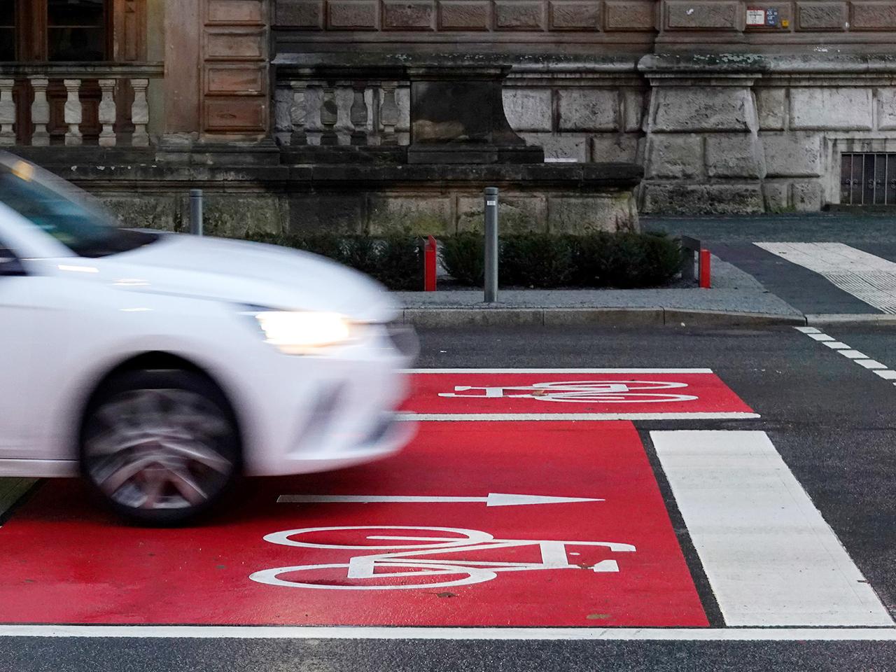 Auf einer Straße ist in weißer Farbe ein Fahrrad und ein Richtungspfeil in Fahrtrichtung auf rotem Grund aufgemalt. Ein weißes Auto fährt über die Kreuzung. 