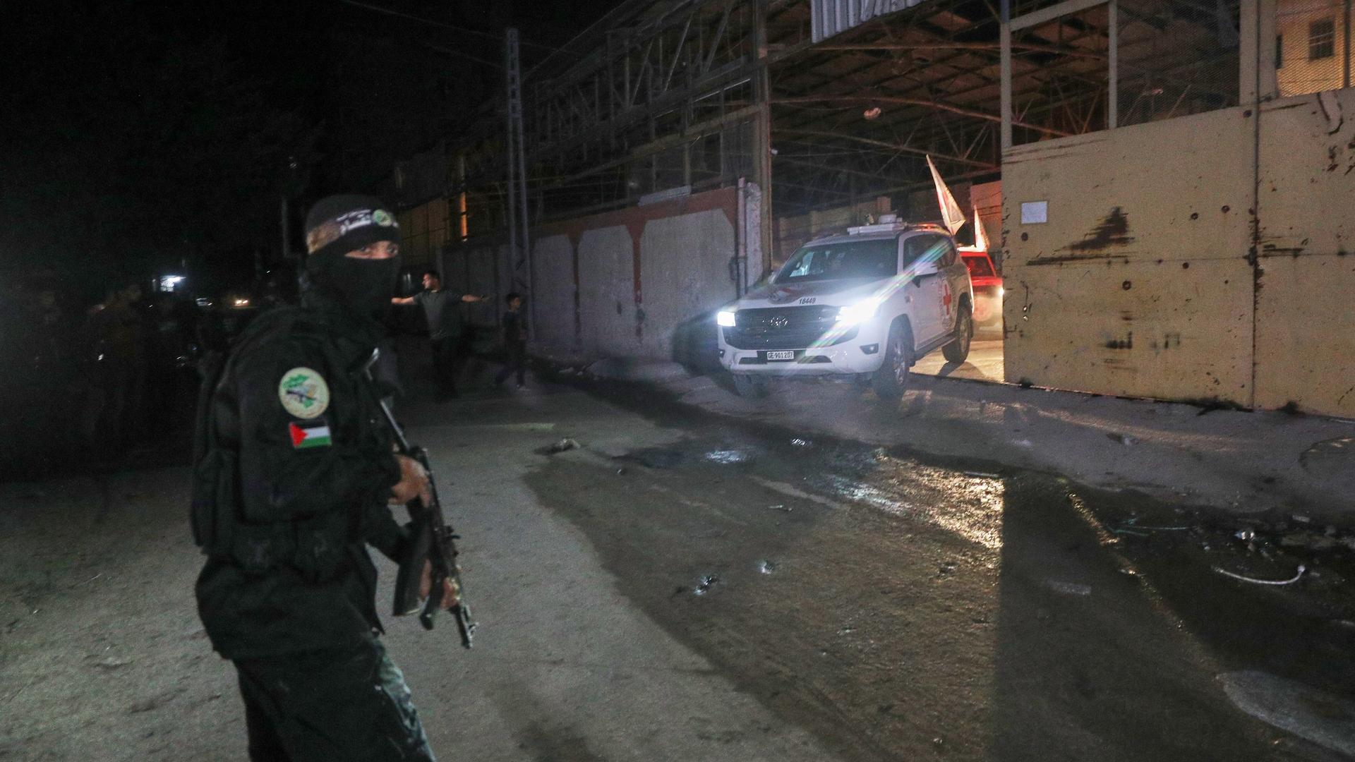 Ein Fahrzeug des Roten Kreuzes, mutmaßlich beladen mit toten Geiseln, verlässt eine Halle in Gaza City; davor steht ein vermummter Kämpfer der al-Qassam-Brigaden Ein Fahrzeug des Roten Kreuzes, mutmaßlich beladen mit toten Geiseln, verlässt eine Halle in Gaza City; davor steht ein vermummter Kämpfer der al-Qassam-Brigaden