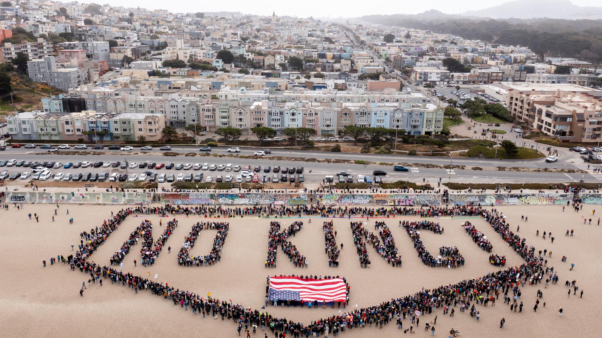 Blick aus großer Höhe auf den Strand. Hunderte Menschen formen den Schriftzug "NO KING!" mit einer Umrandung und einer US-Flagge. Blick aus großer Höhe auf den Strand. Hunderte Menschen formen den Schriftzug "NO KING!" mit einer Umrandung und einer US-Flagge.
