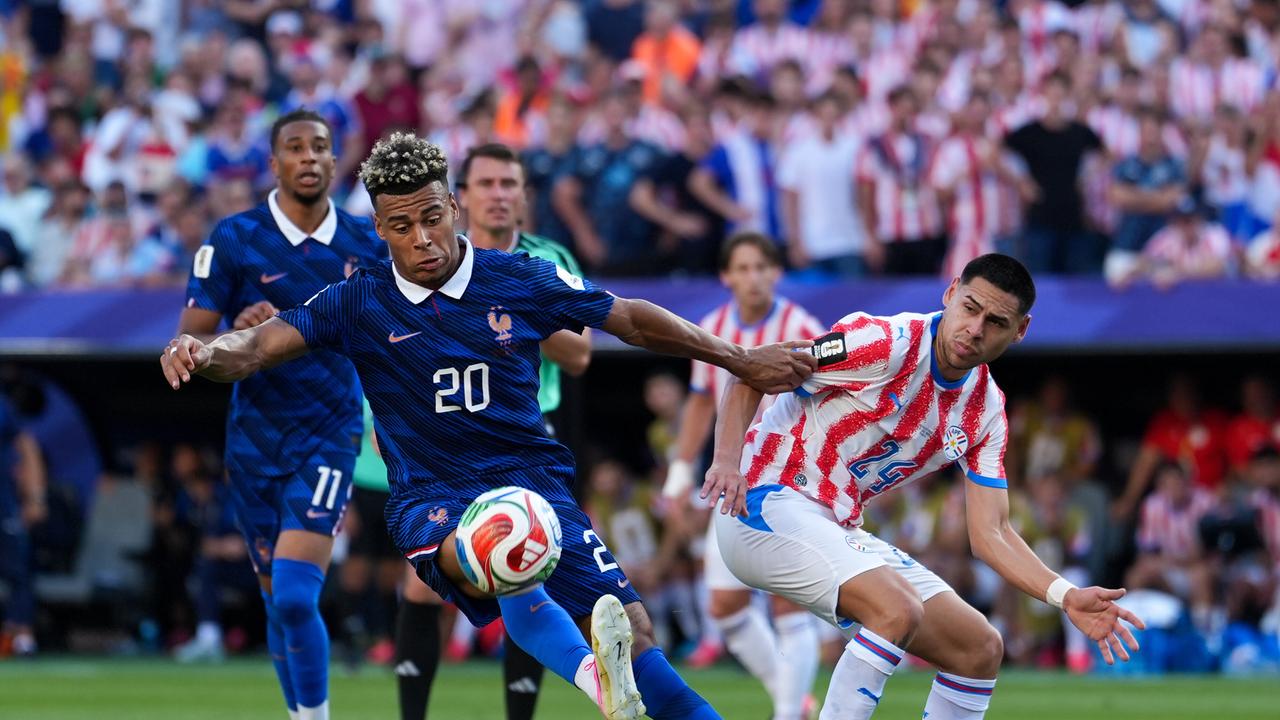 Das Bid zeigt Frankreichs Spieler Desire Doue wie er Gustavo Caballero von der paraguayischen Mannschaft den Ball streitig macht. Doue läuft auf den Betrachter zu, während Caballero zurück bleibt. Im Hintergrund jubelnde Fans.