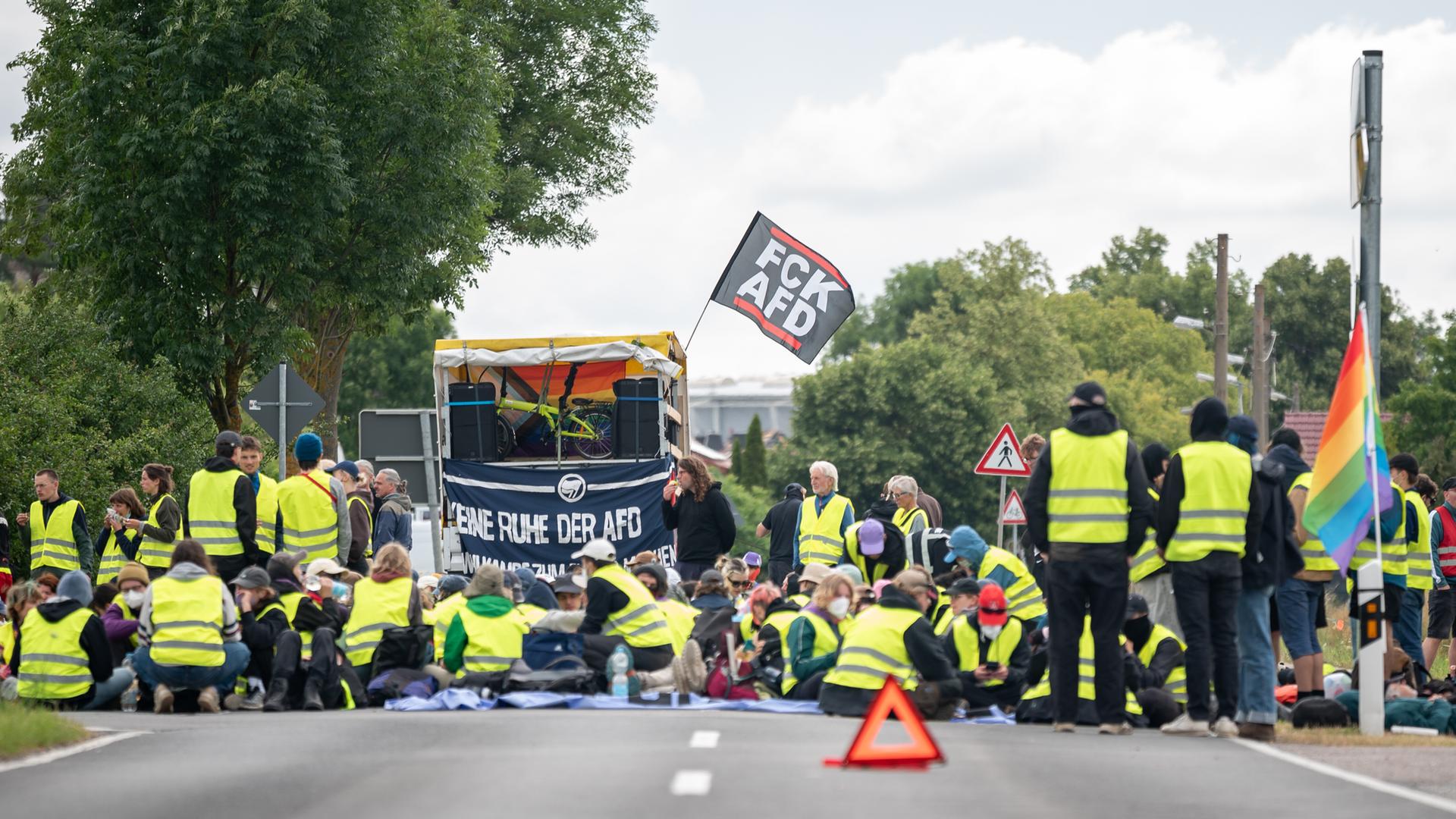 Bei einer Strassenblockade des Bündnis "Widersetzen" in Erfurt sitzen Demonstrierende in gelben Warnwesten auf der Straße. Hinter ihnen ist eine kleine Bühne aufgebaut, an der eine Fahne mit der Aufschrift "FCK AFD" befestigt ist. Bei einer Strassenblockade des Bündnis "Widersetzen" in Erfurt sitzen Demonstrierende in gelben Warnwesten auf der Straße. Hinter ihnen ist eine kleine Bühne aufgebaut, an der eine Fahne mit der Aufschrift "FCK AFD" befestigt ist.
