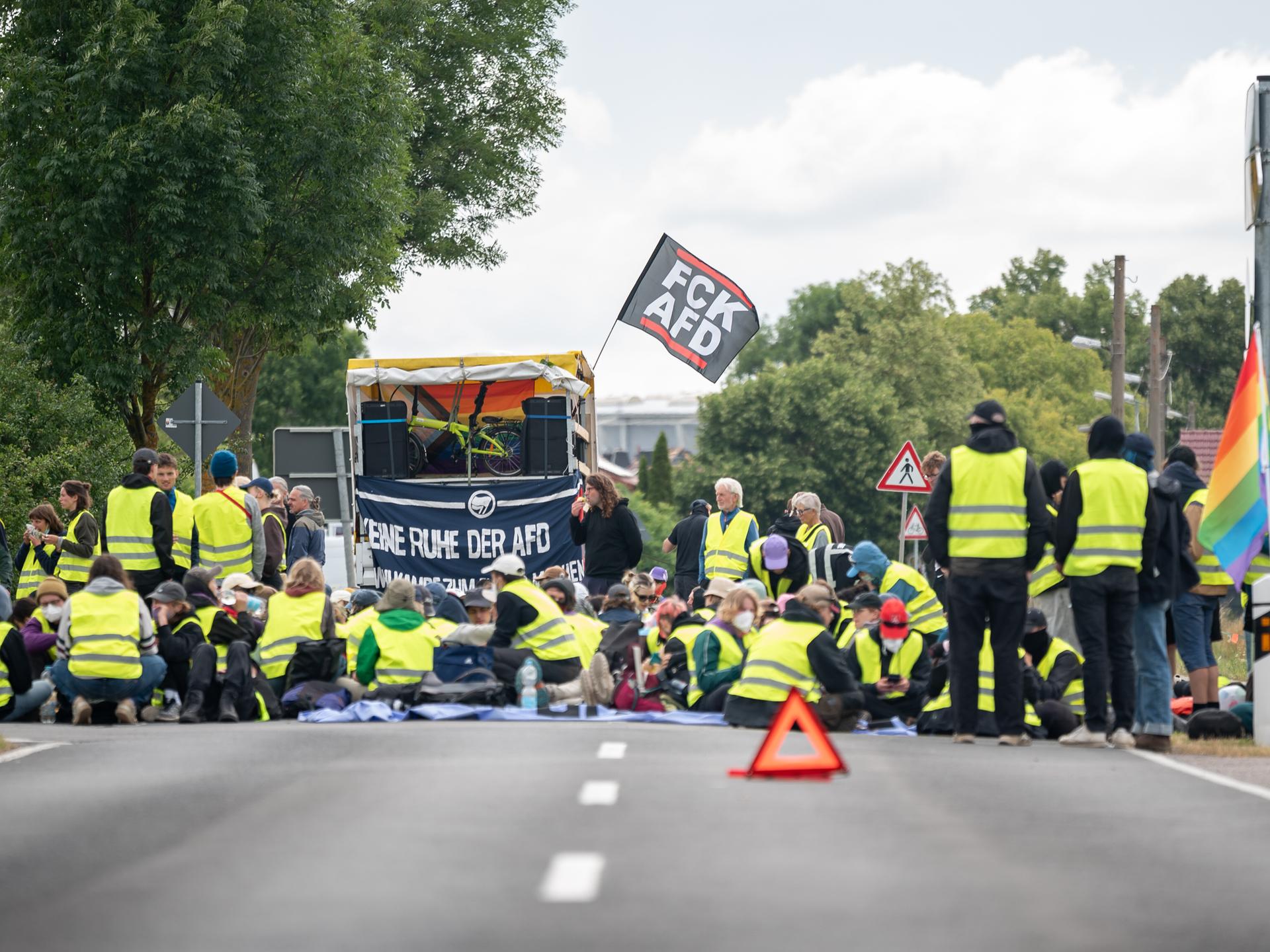 Bei einer Strassenblockade des Bündnis "Widersetzen" in Erfurt sitzen Demonstrierende in gelben Warnwesten auf der Straße. Hinter ihnen ist eine kleine Bühne aufgebaut, an der eine Fahne mit der Aufschrift "FCK AFD" befestigt ist. Bei einer Strassenblockade des Bündnis "Widersetzen" in Erfurt sitzen Demonstrierende in gelben Warnwesten auf der Straße. Hinter ihnen ist eine kleine Bühne aufgebaut, an der eine Fahne mit der Aufschrift "FCK AFD" befestigt ist.