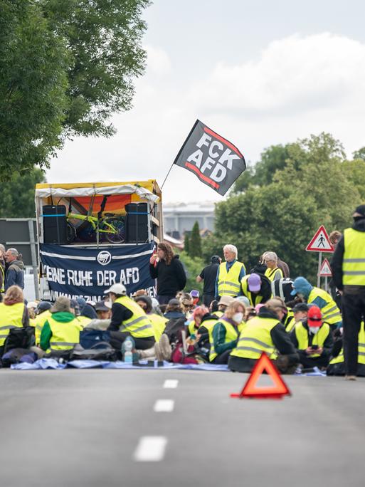 Bei einer Strassenblockade des Bündnis "Widersetzen" in Erfurt sitzen Demonstrierende in gelben Warnwesten auf der Straße. Hinter ihnen ist eine kleine Bühne aufgebaut, an der eine Fahne mit der Aufschrift "FCK AFD" befestigt ist. Bei einer Strassenblockade des Bündnis "Widersetzen" in Erfurt sitzen Demonstrierende in gelben Warnwesten auf der Straße. Hinter ihnen ist eine kleine Bühne aufgebaut, an der eine Fahne mit der Aufschrift "FCK AFD" befestigt ist.