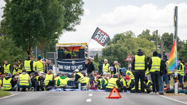 Bei einer Strassenblockade des Bündnis "Widersetzen" in Erfurt sitzen Demonstrierende in gelben Warnwesten auf der Straße. Hinter ihnen ist eine kleine Bühne aufgebaut, an der eine Fahne mit der Aufschrift "FCK AFD" befestigt ist.