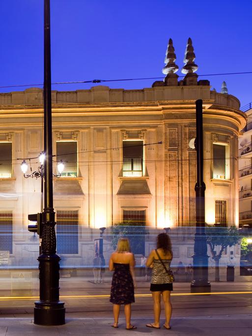 Zwei junge Frauen stehen am Abend in Sevilla vor einer vorbeifahrenden Straßenbahn, im Hintergrund ist ein erleuchtetes Gebäude zu sehen.