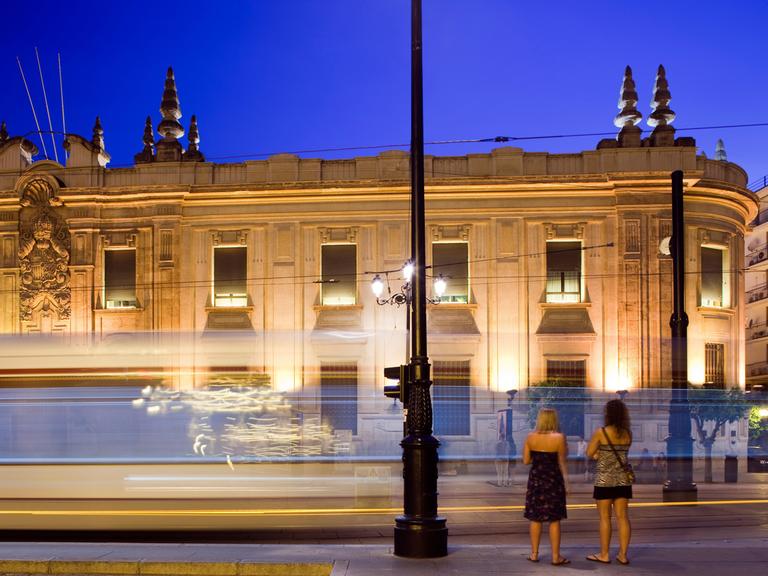 Zwei junge Frauen stehen am Abend in Sevilla vor einer vorbeifahrenden Straßenbahn, im Hintergrund ist ein erleuchtetes Gebäude zu sehen.