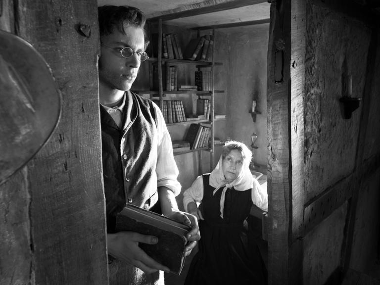 Schwarzweiß-Aufnahme eines Bauernzimmers des 19. Jahrhunderts: Ein junger Mann steht an der Tür, durch die wir eine ältere Frau am Tisch sitzen sehen. Der Gesamteindruck des Bildes ist beengt.