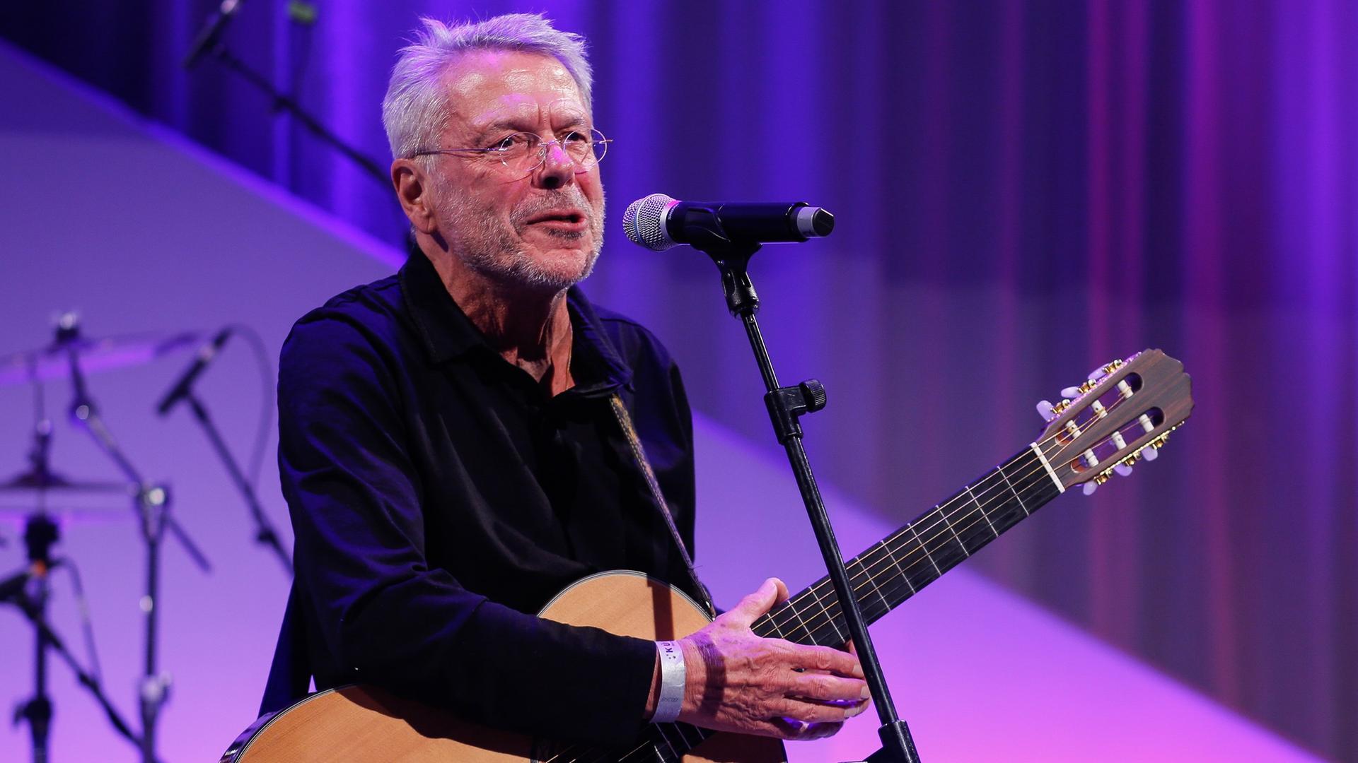 Reinhard Mey steht vor lila-violettem Hintergrund mit seiner Gitarre auf der Bühne. Reinhard Mey steht vor lila-violettem Hintergrund mit seiner Gitarre auf der Bühne.