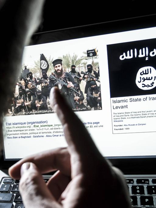Ein Mann sitzt vor einem Computerbildschirm, auf dem eine offensichtlich islamistische Gruppe bärtiger Männer zu sehen ist, die Maschinengewehre in der Hand halten. Daneben sieht man die Flagge vom "Islamistischen Staat". Ein Mann sitzt vor einem Computerbildschirm, auf dem eine offensichtlich islamistische Gruppe bärtiger Männer zu sehen ist, die Maschinengewehre in der Hand halten. Daneben sieht man die Flagge vom "Islamistischen Staat".
